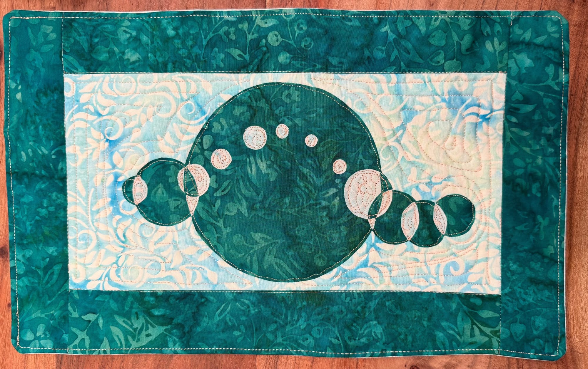 A quilt of overlapping circles with turquoise and blue/white that forms a sine wave flowing down with various sized circles